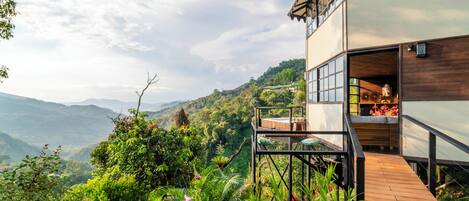 A modern two-story building with a wooden deck and glass walls, overlooking a lush valley.