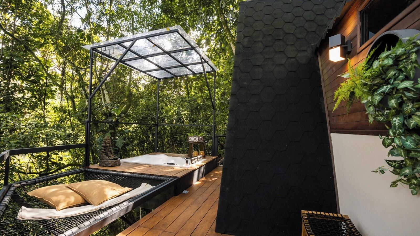 Una terraza de madera con una hamaca, un jacuzzi y una zona de estar con paredes de cristal, rodeada de exuberante vegetación.