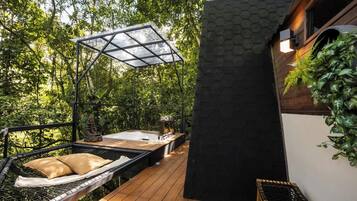 Una terraza de madera con una hamaca, un jacuzzi y una zona de estar con paredes de cristal, rodeada de exuberante vegetación.