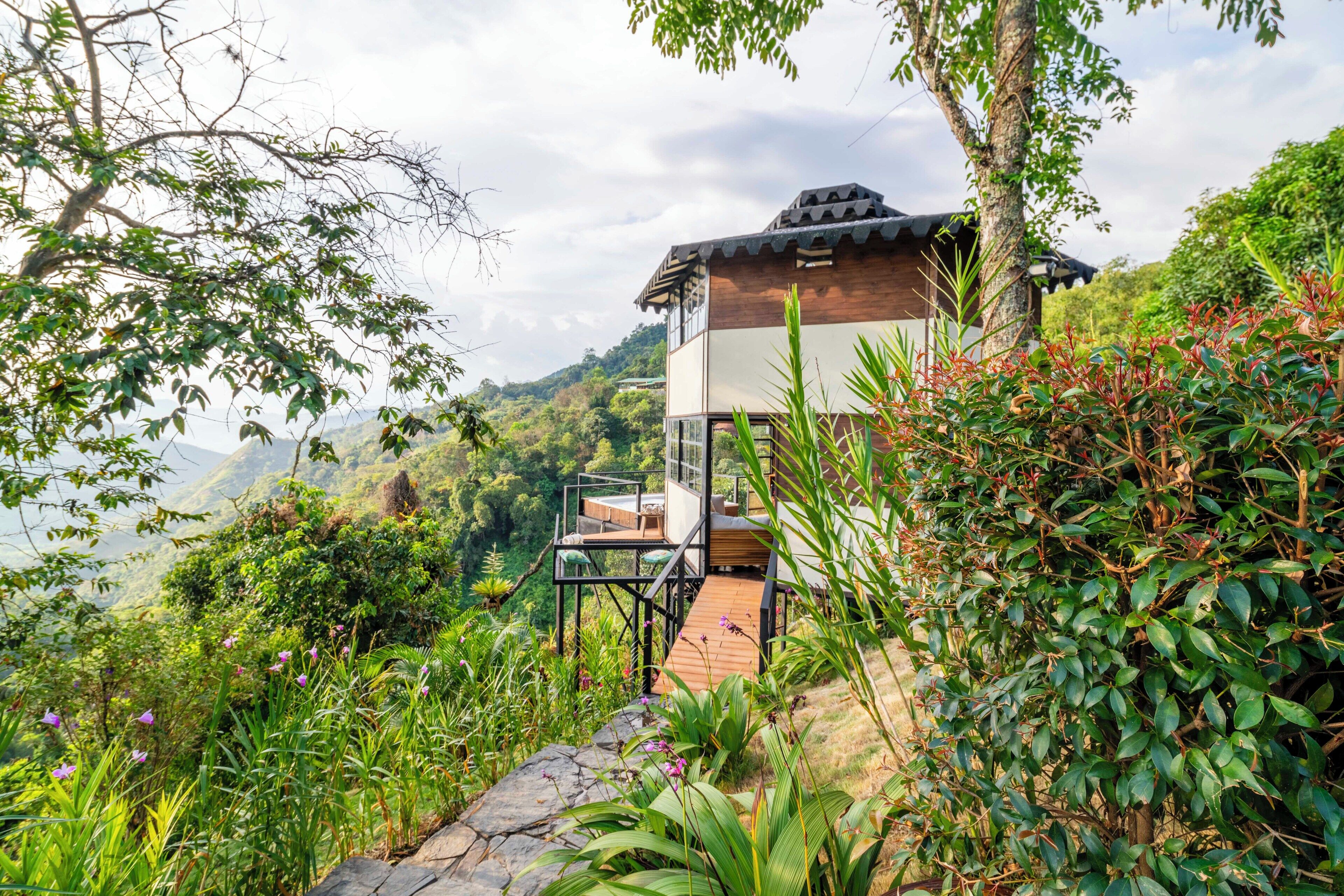 A traditional wooden house with a balcony, surrounded by lush greenery and a mountainous backdrop.