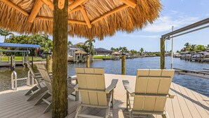 A wooden deck with lounge chairs and a thatched roof structure overlooking a body of water with buildings and palm trees in the background. - What a View -Gulf Access, Southern Exposure, Pool, Ping-Pong-Waterside Oasis - Roelens (Cape Coral)
