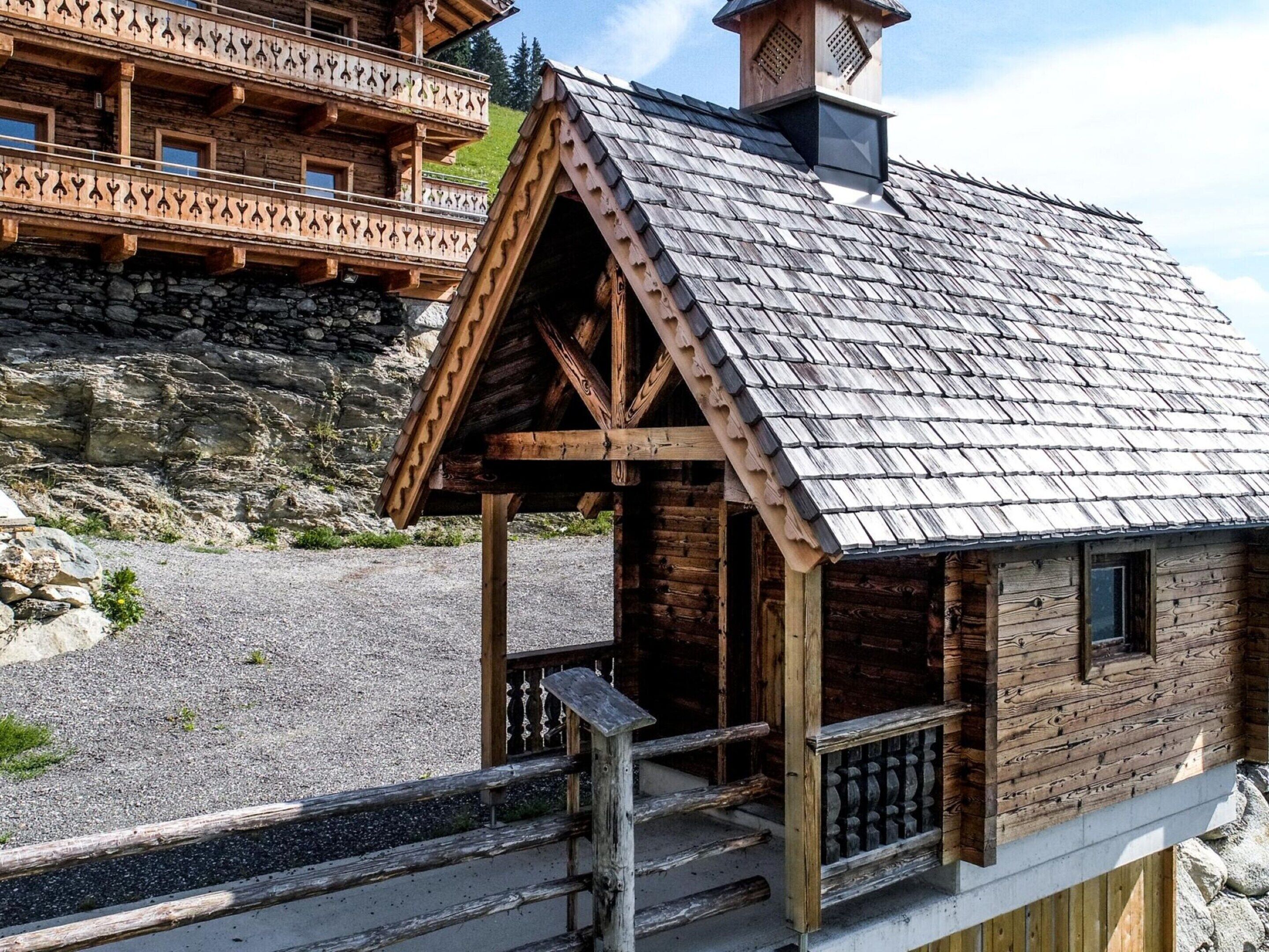 Un chalet de madera con techo a dos aguas, un balcón con barandilla ornamentada y una pared de piedra al fondo.