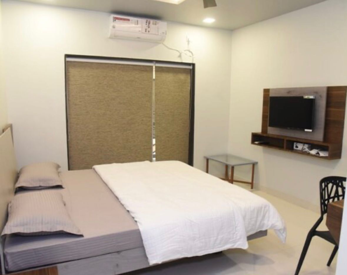 Un dormitorio con una cama, un televisor montado en la pared, una mesita pequeña y una silla.