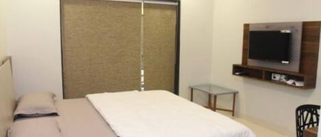 Un dormitorio con una cama, un televisor montado en la pared, una mesita pequeña y una silla.