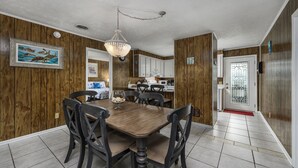 A dining area with a wooden table and chairs, a chandelier, and a kitchen in the background.