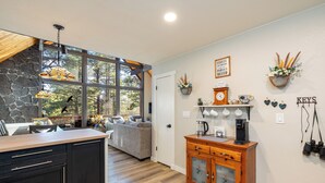 A kitchen with a stone wall, wooden cabinets, a coffee station, and a view through large windows. - Relaxing A-Frame with view of the Pines (Flagstaff)