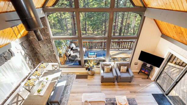 A spacious living area with a large window overlooking a deck, a fireplace, and a flat-screen TV. - Relaxing A-Frame with view of the Pines (Flagstaff)