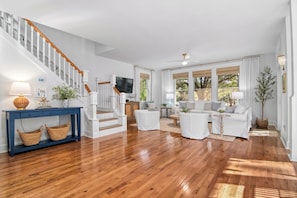 A spacious living room with a wooden floor, white furniture, a staircase, and large windows.