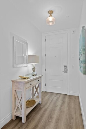 Un couloir avec une porte blanche, une console en bois surmontée d’une lampe, un miroir et un petit objet décoratif.