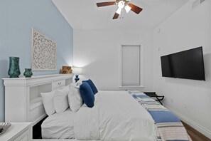 Une chambre à coucher comprenant un lit, un téléviseur, un ventilateur de plafond, une fenêtre et un élément décoratif mural.