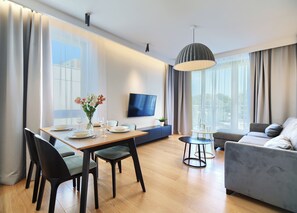 A modern living room with a dining area, a grey sofa, a TV mounted on the wall, and a large window with curtains. - Imperial Apartments - Komandor (Sopot)