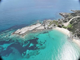 Vista aérea de una zona costera con aguas turquesas cristalinas, terreno rocoso y una playa estrecha.