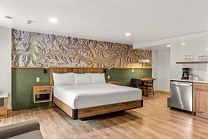 A modern hotel room with a large bed, a wooden headboard, a green accent wall, a small desk, and a kitchenette.