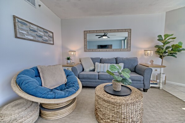 Ein Wohnzimmer mit blauem Sessel, grauem Sofa und einem runden Bistrotisch aus Korbgeflecht.