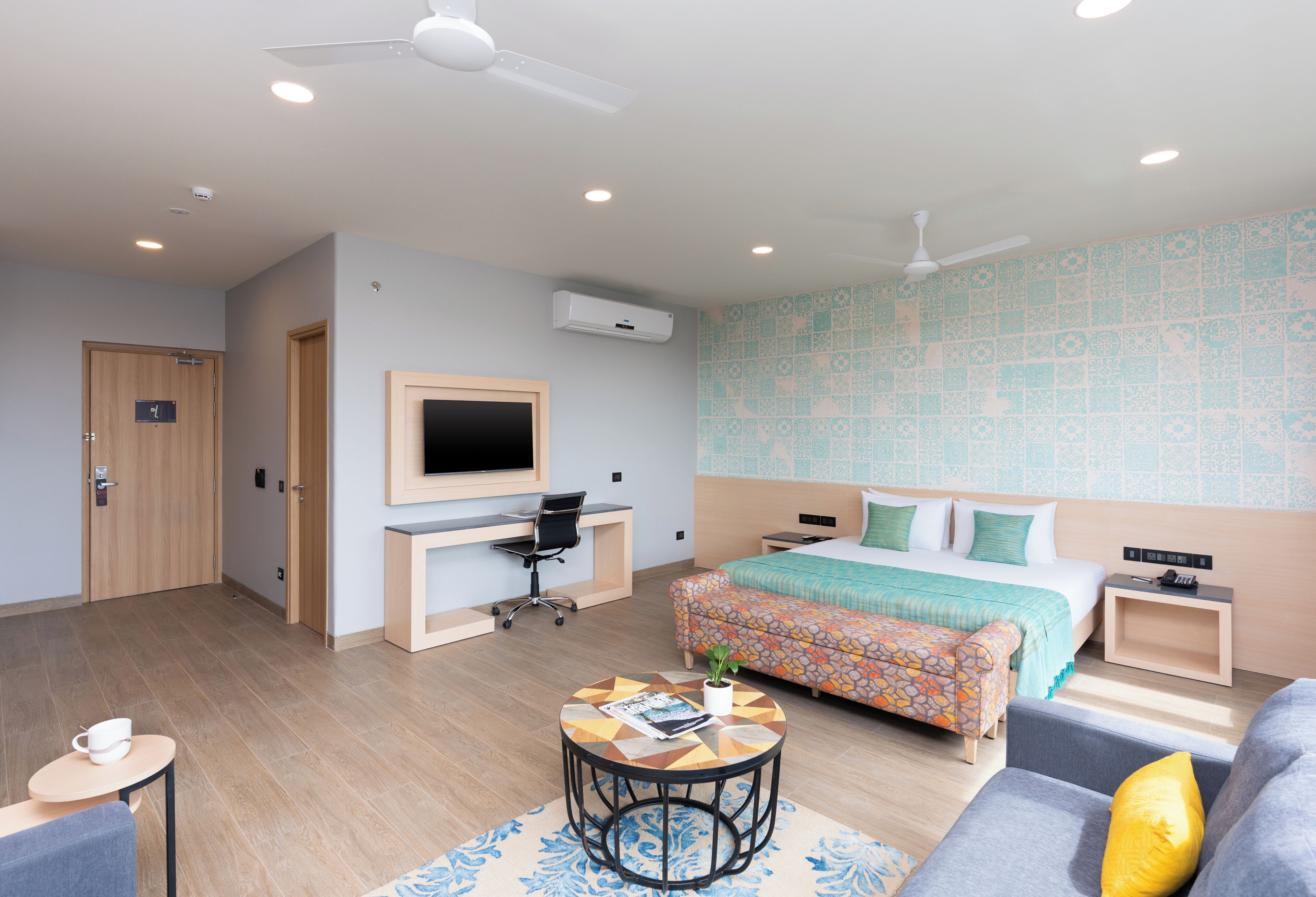 Habitación de hotel con una cama, un escritorio con computadora, una silla, un sofá y una mesita con una planta.