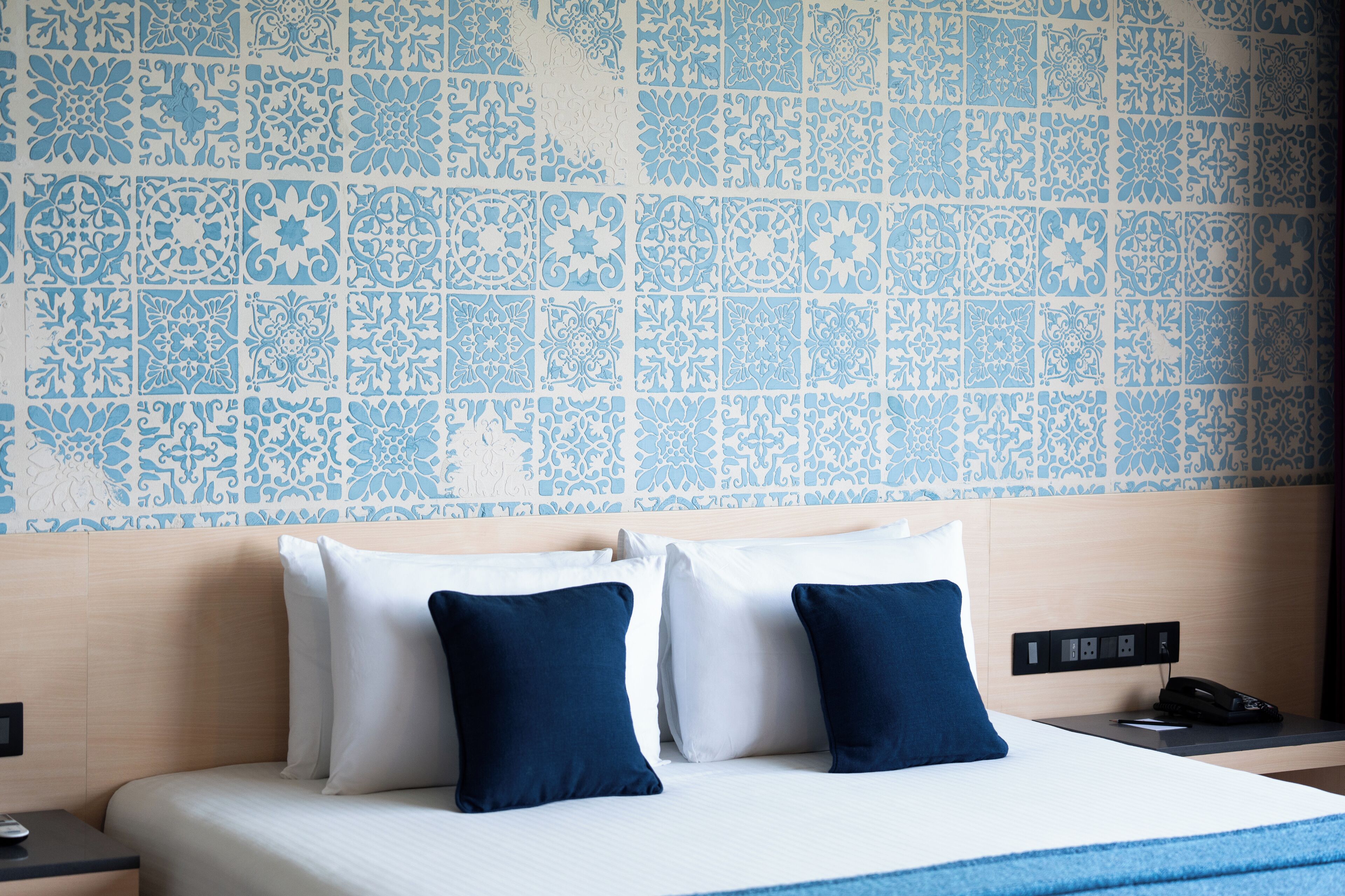 A hotel room with a bed, two pillows, a nightstand, and a wall with blue patterned tiles.