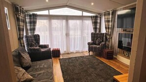 A cozy living room with a large window, a dark rug, and a flat-screen TV. - Tattershall Lakeside Lodge - Tranquil Retreat (Lincoln)