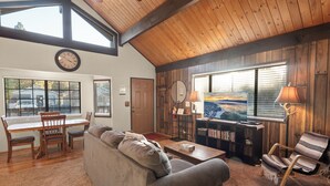 A cozy living room with a wooden ceiling, a large window with a view, a dining area with a table and chairs, and a bookshelf. - Cozy Cabin - Quiet Area, Fireplace, Smart TV, Fenced Yard (Big Bear City)