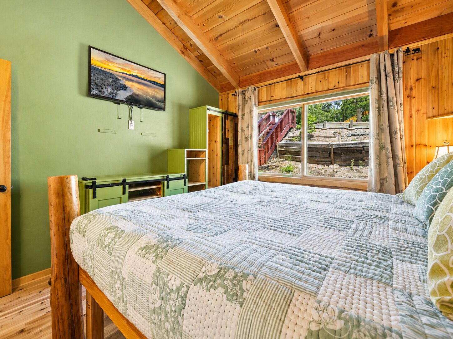 A bedroom with a large bed, a desk, and a window with a view of a wooden deck and trees.