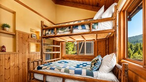 Una cama litera con colcha, paredes de madera, una ventana con vistas y un estante pequeño con una planta.