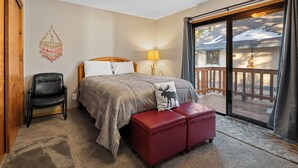A bedroom with a bed, a red ottoman, a chair, a lamp, and a view of a deck.
