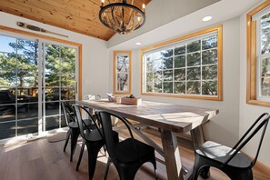 A dining area with a wooden table and chairs, large windows with a view of trees, and a 'Welcome' sign on the wall. - Lucky Paws - Great Location, Hot Tub, Deck, Barbecue (Big Bear City)