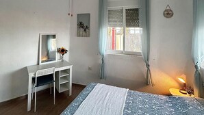 A bedroom with a bed, a desk, a chair, a ceiling fan, a window with curtains, and a wall-mounted lamp.