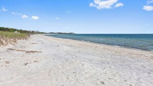 A wide sandy beach with gentle waves, a clear sky, and sparse vegetation. - Private Beach Escape With Sweeping Ocean Views in West Hyannisport RPAM103 (West Hyannisport)