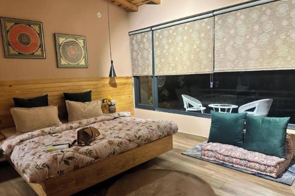 A bedroom with a large bed, a wooden headboard, a window with patterned curtains, and a view of outdoor furniture.