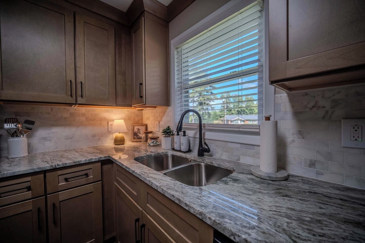 A kitchen with wooden cabinets, a marble countertop, a stainless steel sink, and a window with blinds.
