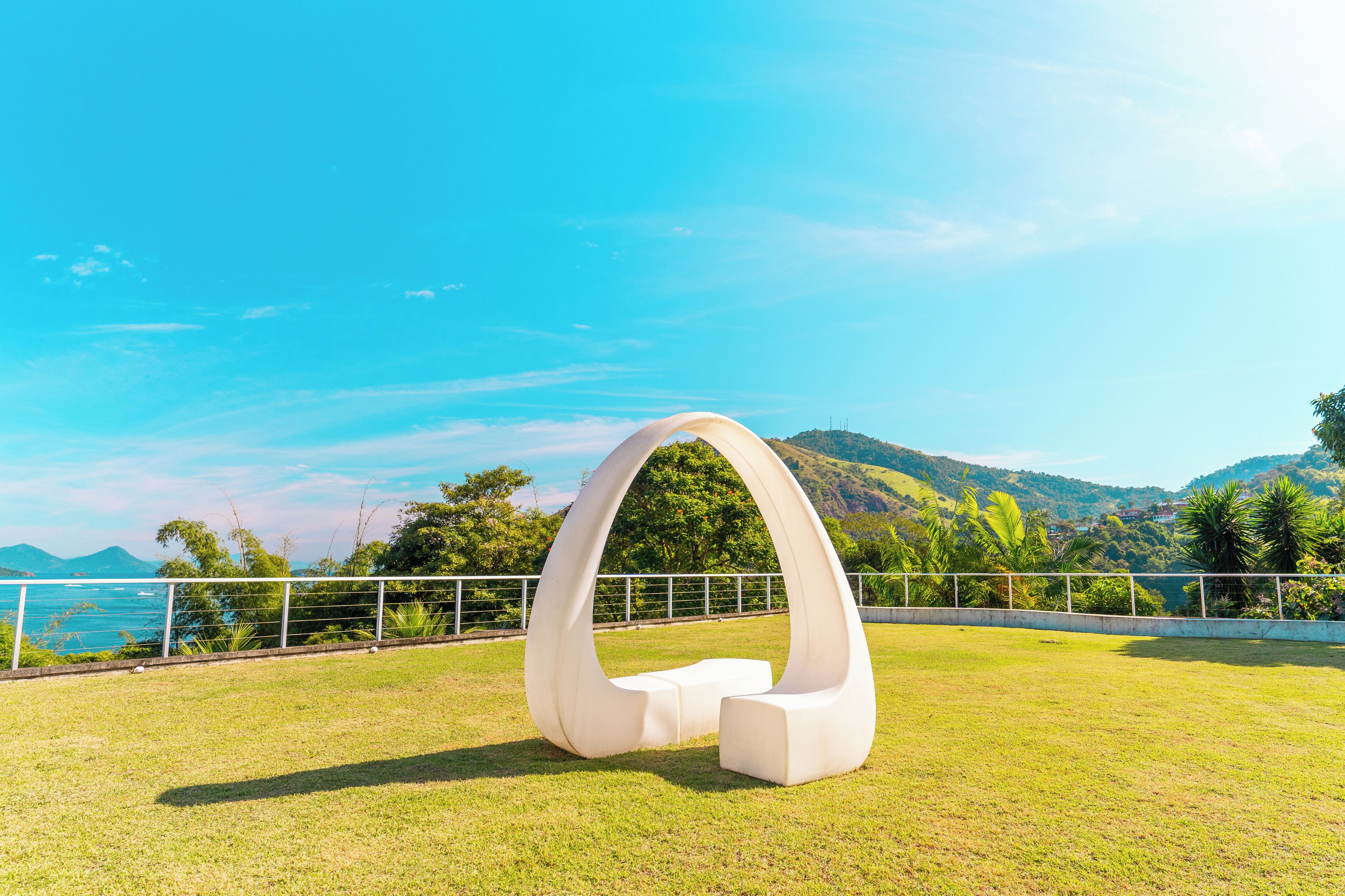 A white, modern sculpture in a grassy field with a clear view of a coastal landscape and hills in the background.
