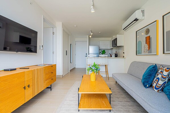 A modern living room with a sofa, coffee table, and TV. There is a kitchen in the background with white cabinets and a microwave.