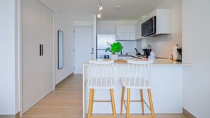 A modern kitchen with white cabinetry, a central island with two white bar stools, a microwave, and a potted plant. - w Majestic 1BR w Stunning Balcony View (Panama City)