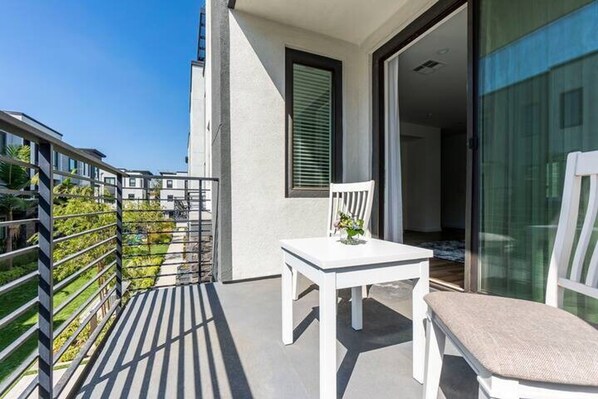 A balcony with a white table and chair, a glass door, and a view of the greenery and neighboring buildings. - Private Room With Shared Bathroom (Costa Mesa)