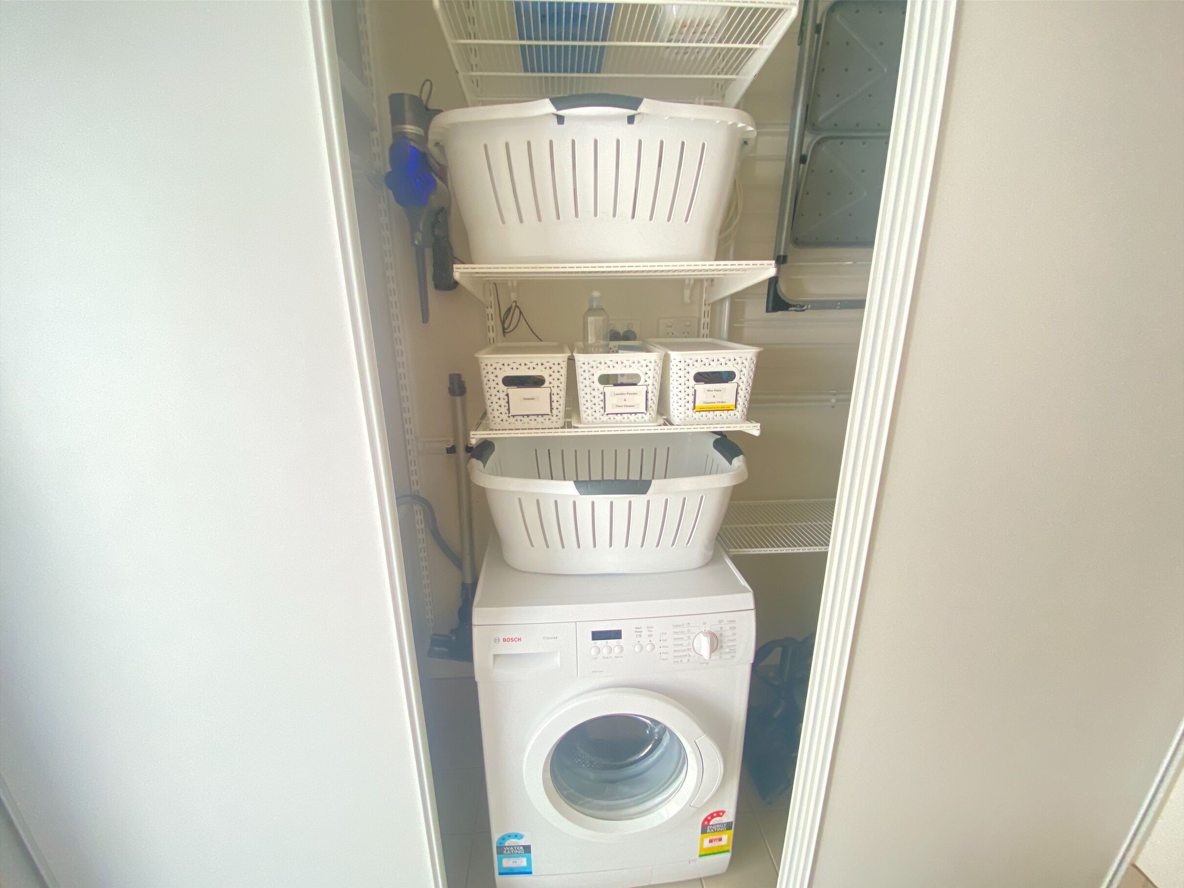 Un cuarto de lavandería con una lavadora Bosch, cestos blancos apilados y estantes con útiles de limpieza.