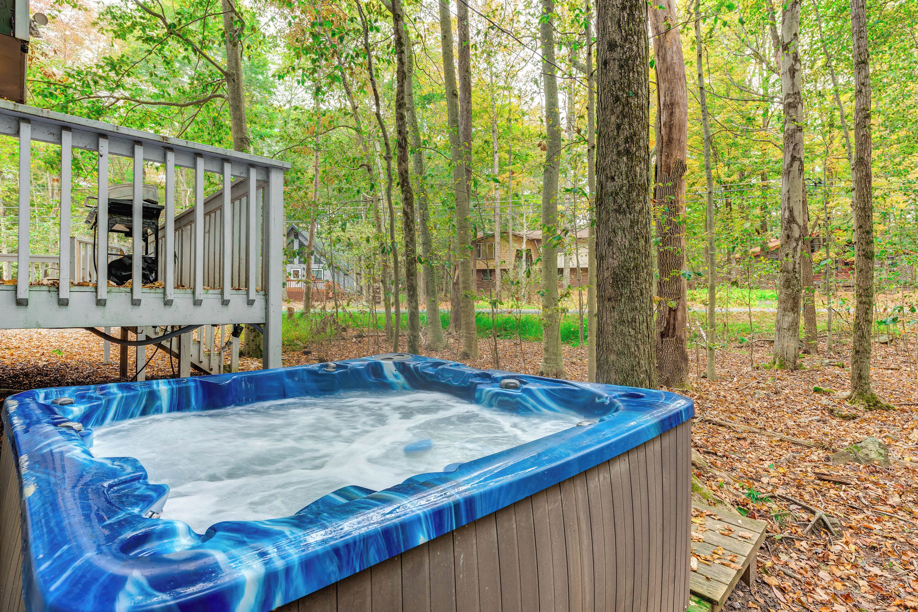 Un bain à remous situé dans un espace boisé, avec une terrasse en bois et une vue sur une maison en arrière-plan.