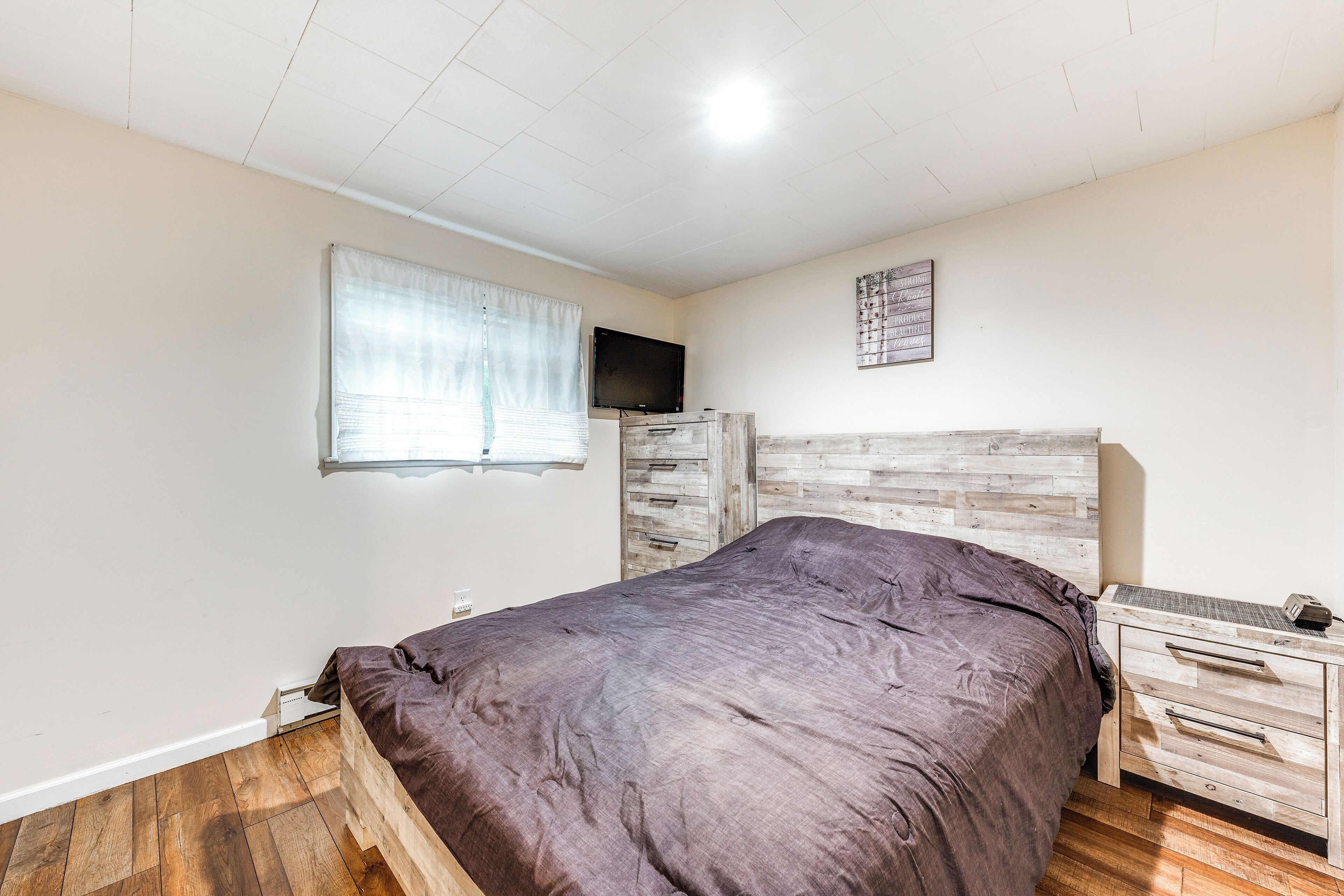 Quarto com cabeceira de madeira, cama com edredom escuro, mesa de cabeceira de madeira com gavetas, televisão de tela plana na parede e janela com cortinas leves.