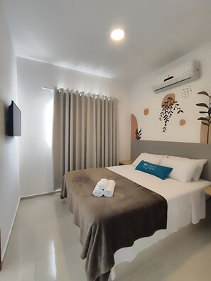 A hotel room with a bed, a flat-screen TV mounted on the wall, a window with curtains, and an air conditioning unit. - Búzios Suíte Home (Búzios)