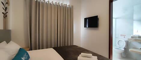 A hotel room with a bed, a TV mounted on the wall, a bathroom visible through an open door, and a towel on the bed.