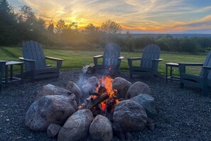 Een vuurkorf met vier Adirondack-stoelen eromheen, gelegen op een grindpad met een zonsondergang op de achtergrond.