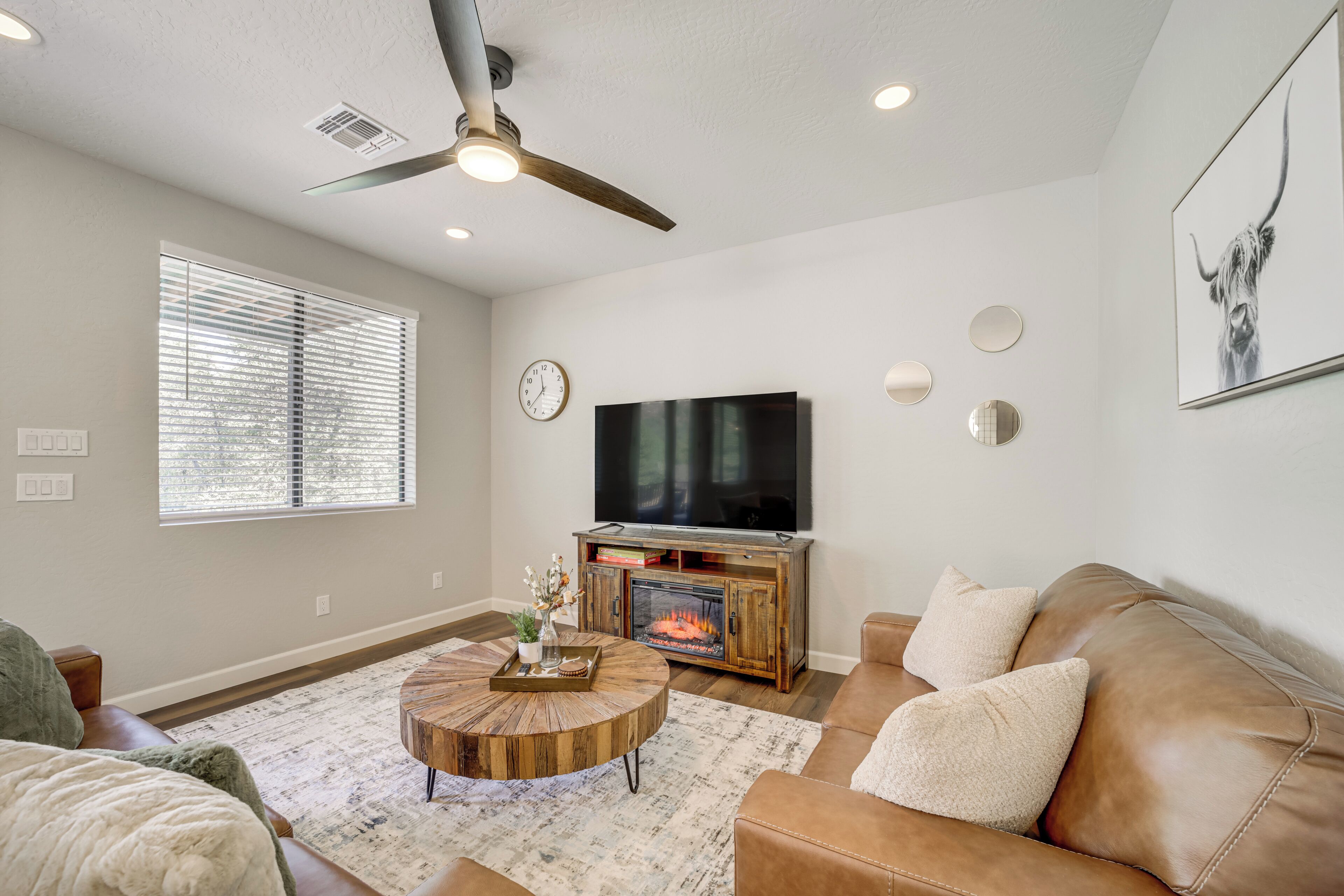 A living room with a brown leather couch, a round coffee table, a fireplace, a flat-screen TV, and a ceiling fan.