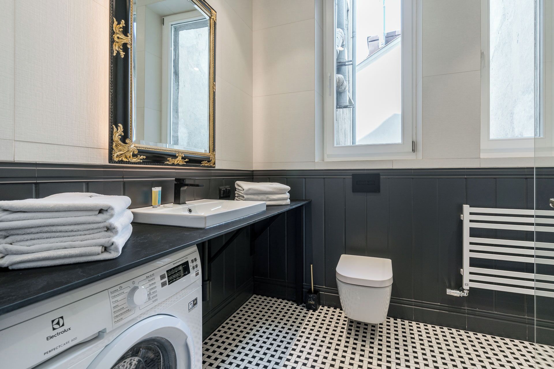 A modern bathroom with a black vanity, white sink, and a large mirror. There is a washing machine and a toilet.