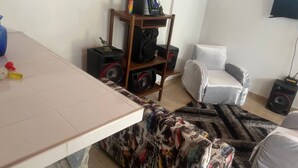 A living room with a white sofa, a coffee table, a patterned ottoman, a wooden shelf with speakers, and a television on the wall. - Country apartment (Ibague)