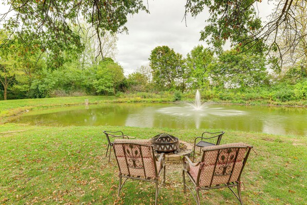 Une table de pique-nique avec des chaises et un foyer, dans un parc doté d’un étang et d’une fontaine.