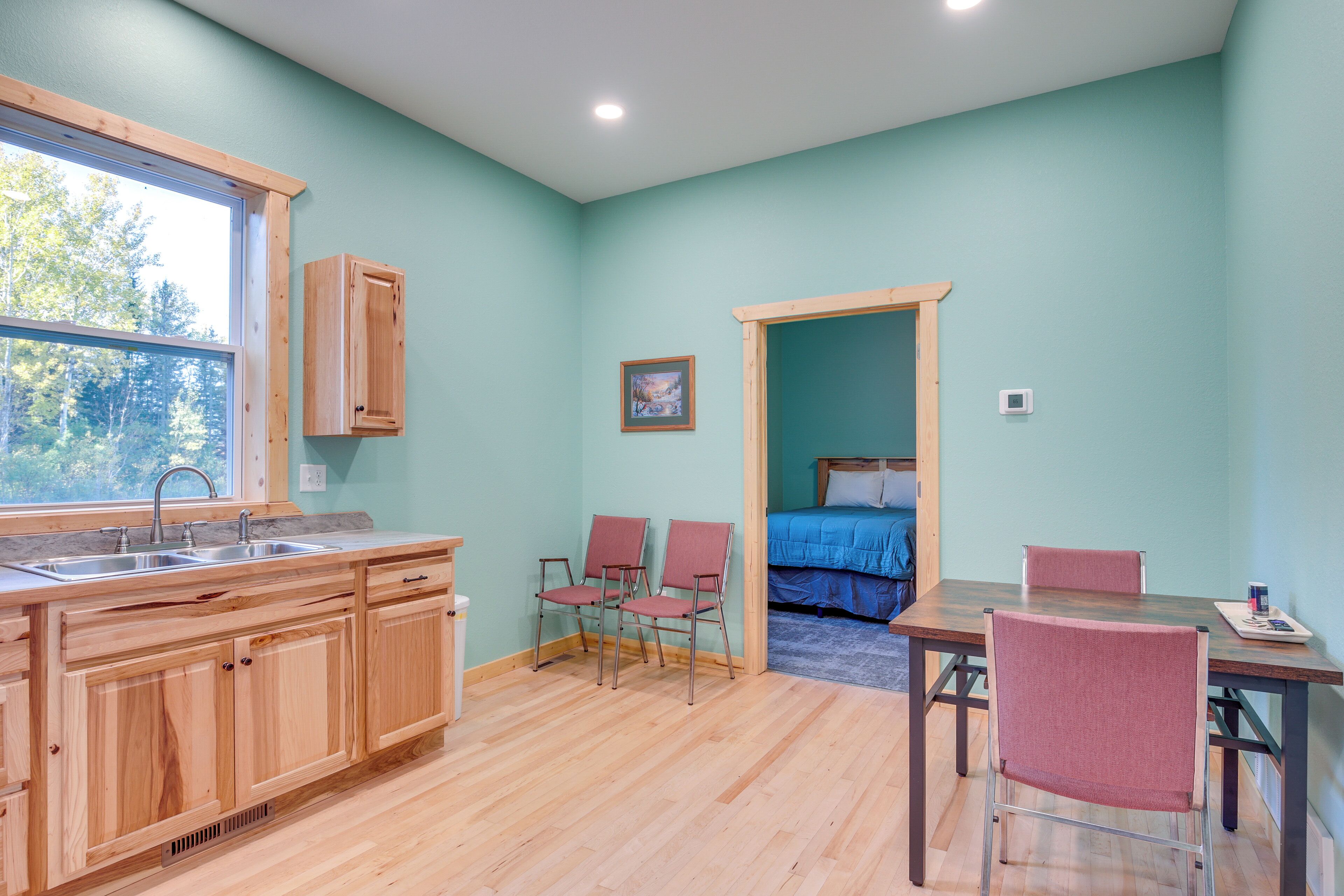 Una habitación con cocina de madera, una mesa de comedor con sillas, una cama con colcha azul y una ventana con vista a árboles.