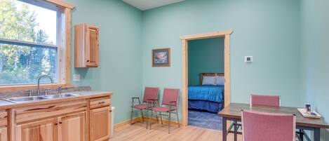 Una habitación con cocina de madera, una mesa de comedor con sillas, una cama con colcha azul y una ventana con vista a árboles.