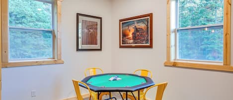 A room with a green pool table, yellow chairs, framed artwork on the walls, and large windows with a view of trees.