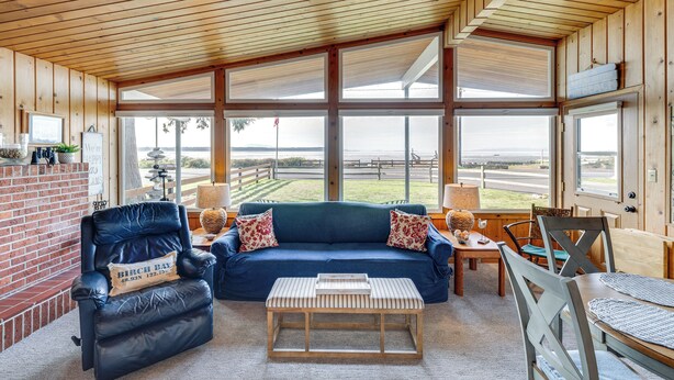 Una habitación acogedora con un sofá azul, un sillón de cuero y una mesa de centro de madera. Amplias ventanas ofrecen vistas al exterior.