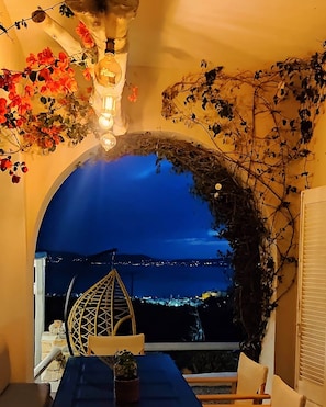 A cozy dining area with a view of the sea at night, featuring a table, chairs, and a decorative archway with plants. - Zephyrus Sunset House (NAXOS)