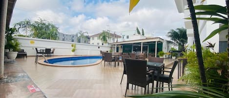 Zona de piscina exterior con suelo de baldosas, rodeada de mobiliario de mimbre y plantas en macetas.